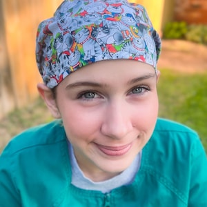 May include: A woman wearing a teal scrub top with a grey and white patterned surgical cap featuring a cat design.