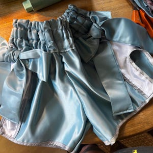 May include: Light blue satin shorts featuring a high-waisted, gathered elastic waistband with a decorative side bow. The shorts have a smooth, reflective finish and are trimmed with a silver-toned metallic border along the leg openings.