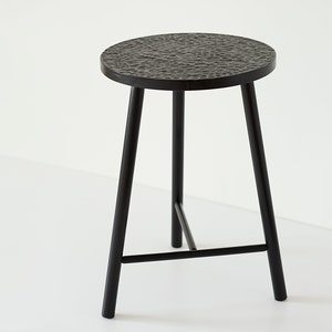 May include: A black metal stool with a round, textured top and three legs. The stool has a simple, modern design.
