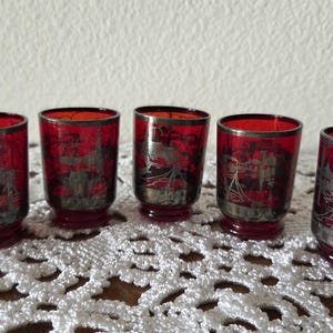 May include: Five vintage red glass shot glasses with silver rims and etched designs. The glasses have a short, rounded base and a cylindrical shape. The etched design appears to be a cityscape or architectural scene.