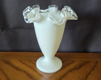 Può includere: Un vaso di vetro bianco con bordo a balze. Il vaso ha una forma svasata e una base a piedistallo. Il bordo è trasparente e il vaso è opaco. Il vaso è su una superficie di legno.
