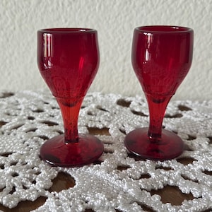 May include: Two ruby red glass cordial glasses with a stemmed base and a rounded bowl. The glasses are displayed on a white, open-weave, lace-like tablecloth. The background is a plain, off-white wall.