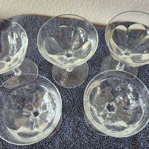 May include: Five clear glass champagne coupes with a diamond pattern on the bowl. The coupes are arranged in a circle on a gray surface.