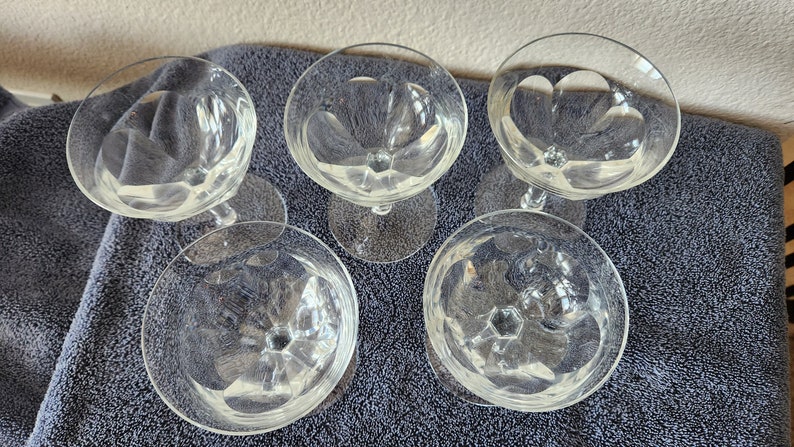 May include: Five clear glass champagne coupes with a diamond pattern on the bowl. The coupes are arranged in a circle on a gray surface.
