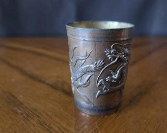 Può includere: Una piccola tazza antica color rame con intricate incisioni a rilievo di draghi. La tazza ha un aspetto leggermente appannato, con un interno color oro. Il disegno del drago avvolge l'esterno della tazza, mostrando squame e dettagli.