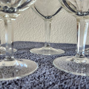 May include: Four clear glass wine glasses with a diamond pattern on the bowl and a long, thin stem. The glasses are sitting on a gray, textured surface.