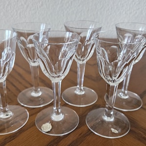 May include: Set of six clear crystal cordial glasses. Each glass features a delicate, etched floral design on the bowl and stem. The glasses are arranged on a wooden surface, showcasing their elegant shape and clarity.