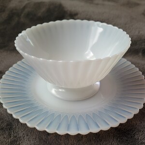Pu&ograve; includere: Una ciotola di vetro opalescente bianco con bordo smerlato poggia su un piattino abbinato. Entrambi i pezzi presentano un design delicato e scanalato.