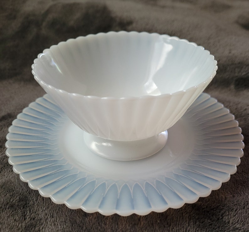 Pu&ograve; includere: Una ciotola di vetro opalescente bianco con bordo smerlato poggia su un piattino abbinato. Entrambi i pezzi presentano un design delicato e scanalato.