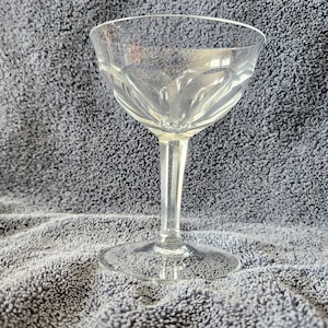 May include: A clear glass champagne coupe with a delicate, fluted design. The coupe is sitting on a gray, textured surface.