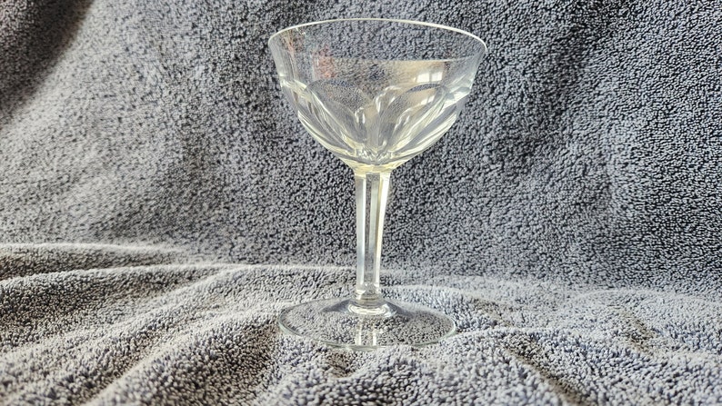 May include: A clear glass champagne coupe with a delicate, fluted design. The coupe is sitting on a gray, textured surface.