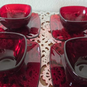 May include: Four matching ruby red glass teacups and saucers. The cups are square with rounded corners and small handles. The saucers are also square. The set is displayed on a white lace tablecloth.