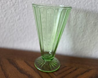 May include: A vintage, light green glass sundae or parfait glass. The glass has a flared, conical shape with vertical ribbed detailing and a circular base. The glass is displayed on a wooden surface against a white wall.