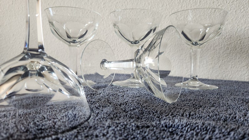 May include: Four clear glass champagne coupes with a cut glass design. The glasses are arranged on a gray textured surface.