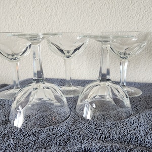 May include: Set of five clear glass champagne coupes with a fluted design. The coupes are arranged in a row on a gray towel.