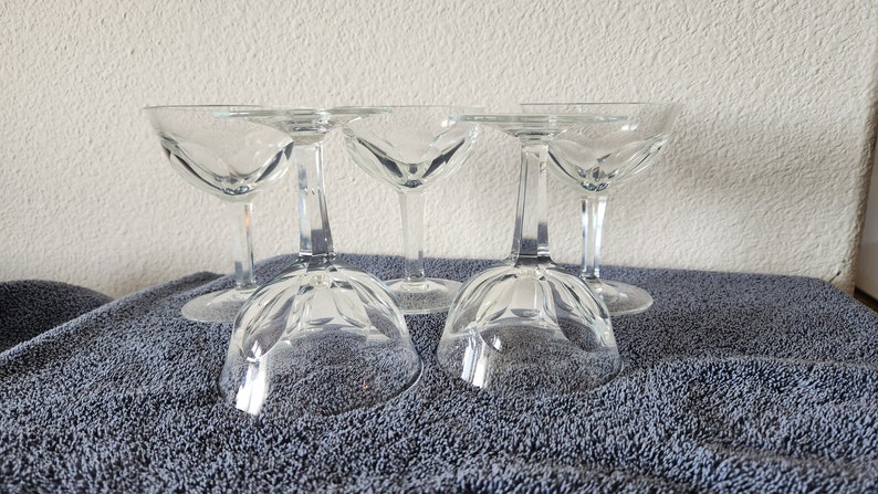 May include: Set of five clear glass champagne coupes with a fluted design. The coupes are arranged in a row on a gray towel.
