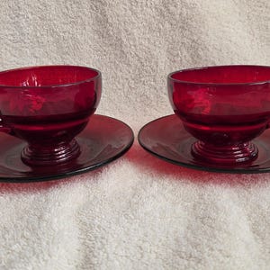 May include: Two ruby red glass teacups and saucers. The cups have a rounded bowl shape with a handle and sit on matching round saucers. The set is made of a transparent, deep red glass, perfect for serving tea or coffee.