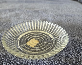 Può includere: Un piatto di vetro trasparente con bordo scanalato e un design a spirale circolare al centro. Il piatto ha una piccola etichetta colorata al centro. Il piatto è appoggiato su una superficie grigia strutturata.