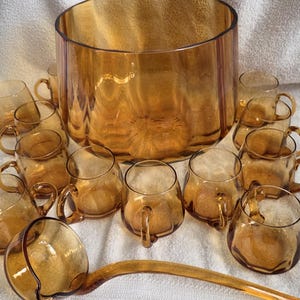 May include: A vintage amber glass punch bowl set. The set includes a large, round punch bowl, a ladle, and several small handled cups. The glassware has a warm, golden hue and is arranged on a white surface.