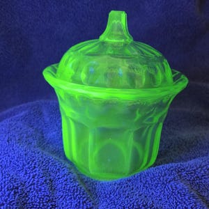 May include: A glowing green glass jar with a lid, set against a blue background. The jar has a decorative, fluted design and a small handle on the lid. The glass appears to be vintage or antique.