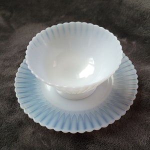 Pu&ograve; includere: Una ciotola in vetro di latte bianco con un bordo a festoni e un piatto abbinato. La ciotola e il piatto hanno una glassa blu iridescente.