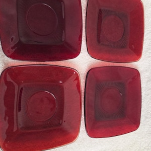 May include: Four square, ruby red glass plates. Each plate has a slightly rounded square shape with a circular indentation in the center. The plates are arranged in a 2x2 grid, showcasing their uniform design and color.
