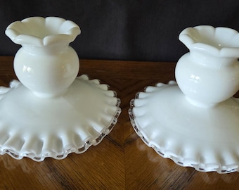 Può includere: Due portacandele in vetro bianco abbinati. Ognuno ha una base a balze e una parte superiore a forma di vaso con un bordo smerlato. I portacandele sono esposti su una superficie di legno.