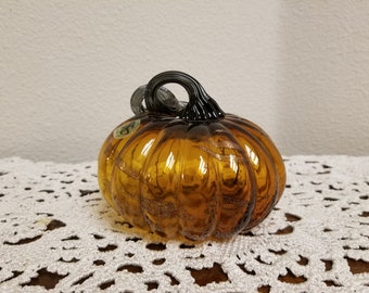 May include: A small amber glass pumpkin with a black glass stem. The pumpkin has a shiny, iridescent finish.