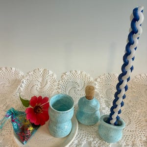 May include: A set of light blue speckled ceramic items including a small plate, a cup, a bottle with a cork stopper, and a small holder containing a tall, spiraled blue and white candle. A small colorful pouch and a red zinnia flower are present.