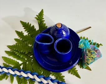 Op de afbeelding: Een kobaltblauwe keramische set met een kleine container met deksel, twee kleine kopjes en een bord. Een gevlochten blauw-witte kaars en een kleine cadeauzak zijn ook aanwezig. De set wordt gepresenteerd op een bed van groene varens.