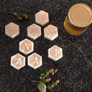 May include: Eight hexagonal shaped soaps with a honeycomb pattern and a bee design. The soaps are arranged in a circle on a dark grey surface. There is a small jar of honey in the background.