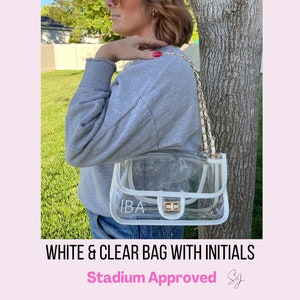 May include: A clear and white plastic bag with a gold chain strap. The bag has a white flap with the initials "IBA" printed on it. The bag is stadium approved.