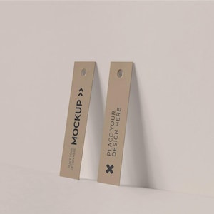 May include: Two brown paper tags with rounded corners and a hole at the top. The tag on the left says "PLACE YOUR DESIGN HERE MOCKUP >>". The tag on the right says "PLACE YOUR DESIGN HERE" with a small black cross.