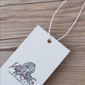 May include: A white rectangular hang tag with a black and white illustration of a woman's hair with pink highlights. The text "Muse Hair Co." is written in black cursive with the tagline "The art of glamorous extensions" in black text below the logo.