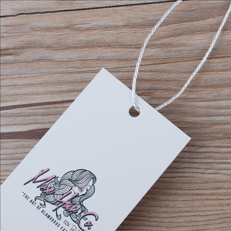 May include: A white rectangular hang tag with a black and white illustration of a woman's hair with pink highlights. The text "Muse Hair Co." is written in black cursive with the tagline "The art of glamorous extensions" in black text below the logo.