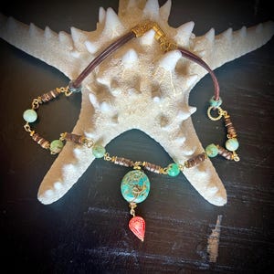 May include: A handcrafted necklace featuring a brown leather cord and gold-toned metal accents. The necklace includes turquoise and green beads, with a central turquoise stone wrapped in gold wire and a red pendant.