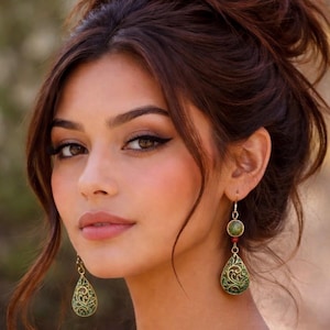 May include: Gold-toned earrings with green and red accents. The earrings feature a teardrop-shaped pendant with an intricate, openwork design. The top of the earrings have a round green stone and a small red bead.