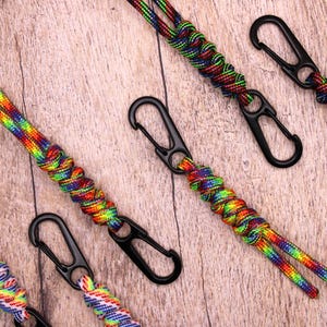 May include: A collection of rainbow-coloured paracord keychains with black metal carabiner clips, arranged on a wooden surface. The keychains display a spectrum of vibrant colours, including red, orange, yellow, green, blue, and purple.