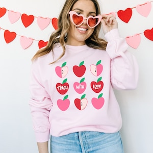 Puede incluir: Sudadera rosa con un gr&aacute;fico de corazones de manzana rojos y rosas. El texto "Teach with Love" est&aacute; impreso en la sudadera.