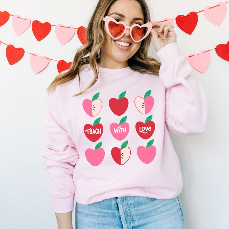 Puede incluir: Sudadera rosa con un gr&aacute;fico de corazones de manzana rojos y rosas. El texto "Teach with Love" est&aacute; impreso en la sudadera.