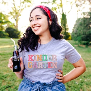 May include: A woman wearing a grey t-shirt with the words "KINDERGARTEN" in colorful letters. She is holding a bottle of Coca-Cola with a straw.