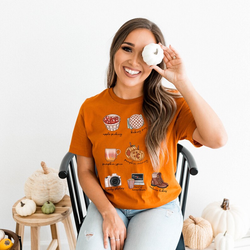 May include: A rust-colored t-shirt with a graphic design of fall activities. The design includes illustrations of apple picking, baking pies, pumpkin spice, pumpkin carving, family photos, movie marathon, and making cookies. The text "apple picking", "baking pies", "pumpkin spice", "pumpkin carving", "family photos", "movie marathon", and "make cookies" is included in the design.