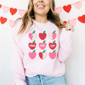 Puede incluir: Sudadera rosa con un dise&ntilde;o gr&aacute;fico de manzanas rojas y rosas con hojas verdes. Las manzanas est&aacute;n dispuestas en una cuadr&iacute;cula y tienen las palabras "Teach with Love" escritas en ellas.