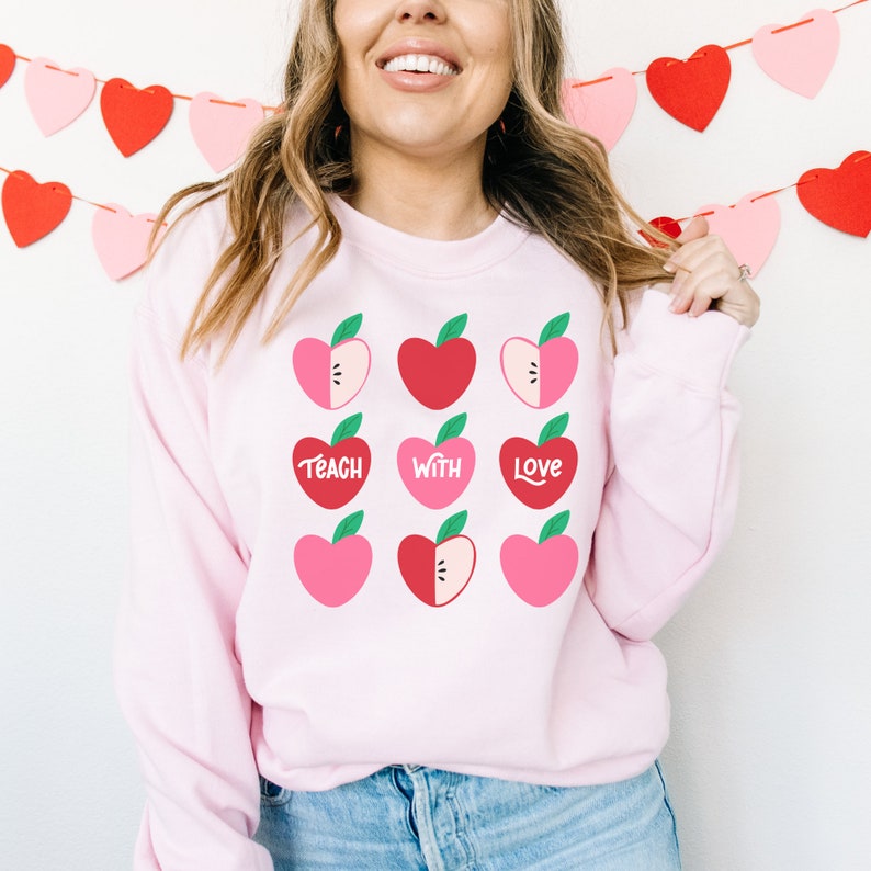 Puede incluir: Sudadera rosa con un dise&ntilde;o gr&aacute;fico de manzanas rojas y rosas con hojas verdes. Las manzanas est&aacute;n dispuestas en una cuadr&iacute;cula y tienen las palabras "Teach with Love" escritas en ellas.