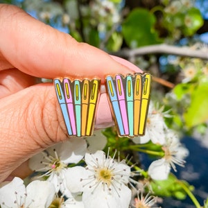 May include: A pair of gold-toned enamel pins featuring a stack of colorful pens. The pens are in shades of blue, pink, yellow, and green.