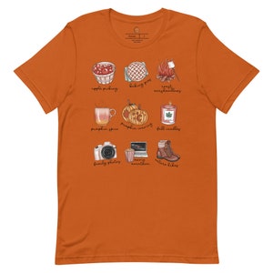 May include: Orange t-shirt with a white graphic of fall activities. The graphic includes images of apple picking, baking pies, roasting marshmallows, pumpkin spice, pumpkin carving, fall candles, family photos, a marathon, and nature hikes.