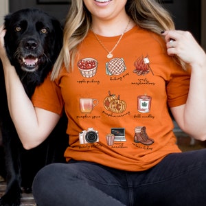 May include: A burnt orange t-shirt with a graphic design of fall activities. The design includes illustrations of apple picking, baking pies, roasting marshmallows, pumpkin spice, pumpkin carving, fall candles, family photos, movie marathon, and nature hikes. The text "apple picking", "baking pies", "roast marshmallows", "pumpkin spice", "pumpkin carving", "fall candles", "family photos", "movie marathon", and "nature hikes" is printed below each illustration.