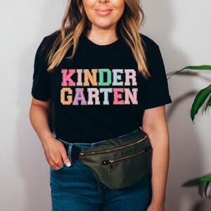 May include: A black t-shirt with the words "KINDERGARTEN" in rainbow colors. The shirt is being worn by a person with a green fanny pack.
