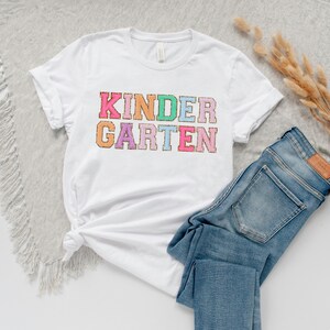 May include: White t-shirt with the text "KINDERGARTEN" in colorful, glittery letters.