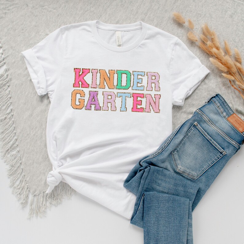 May include: White t-shirt with the text "KINDERGARTEN" in colorful, glittery letters.